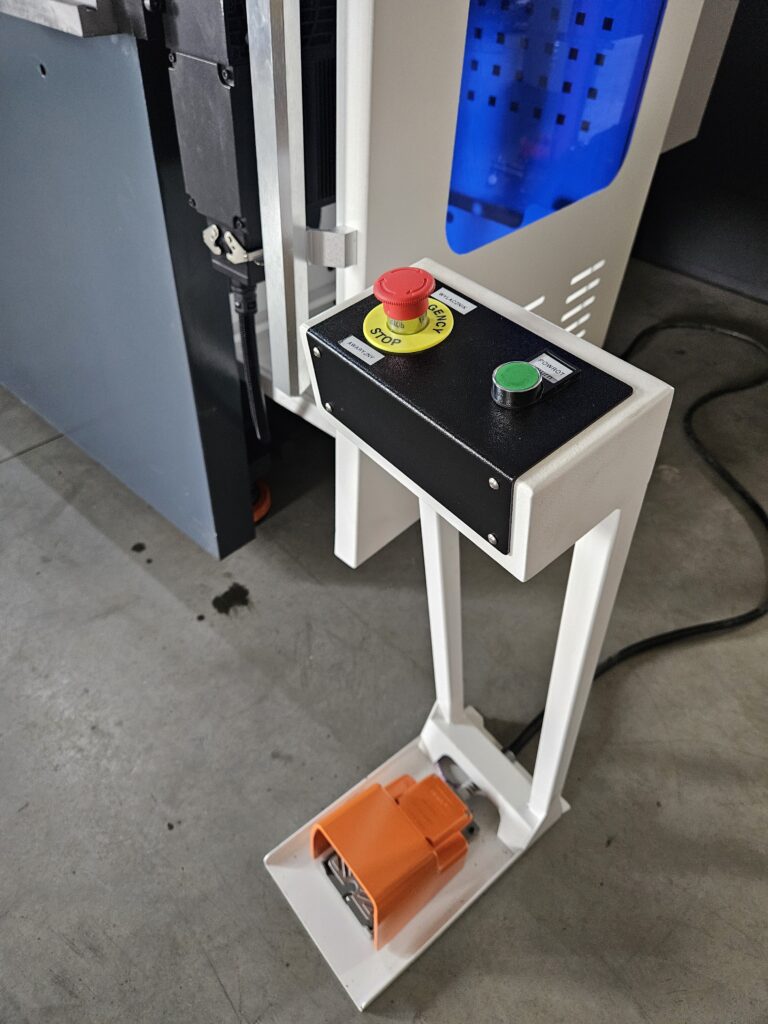 Wyłącznik bezpieczeństwa z czerwonym przyciskiem "EMERGENCY STOP" i zielonym "START" na szarej maszynie.